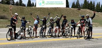 Riders in cycling jerseys raise their arms beside a Grief Center banner on a mountain road
